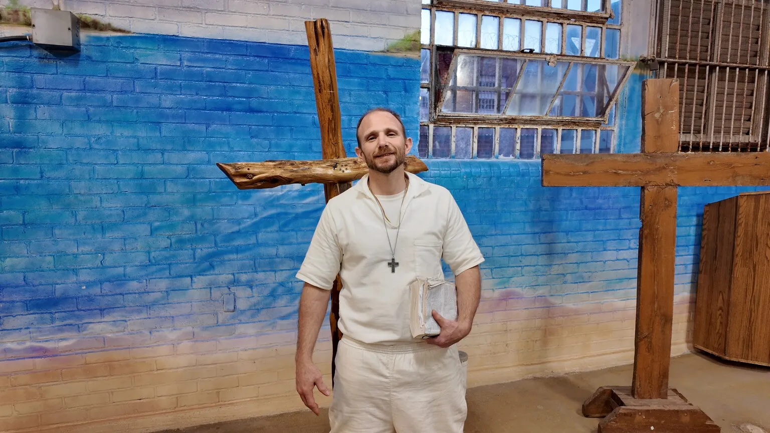 Man in white uniform standing by wooden cross mural