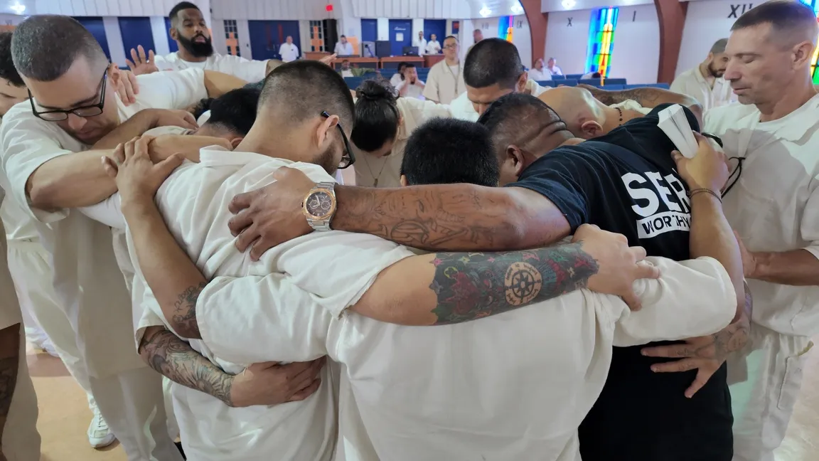 Inmates in white uniforms embracing in group prayer huddle