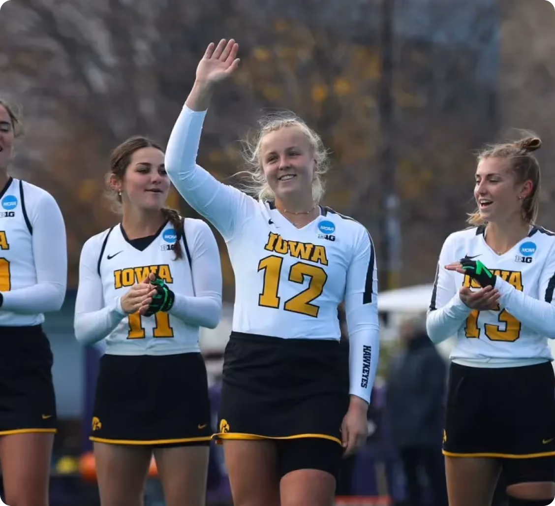 Iowa female athletes in white jerseys and black skirts waving and clapping during an outdoor event.
