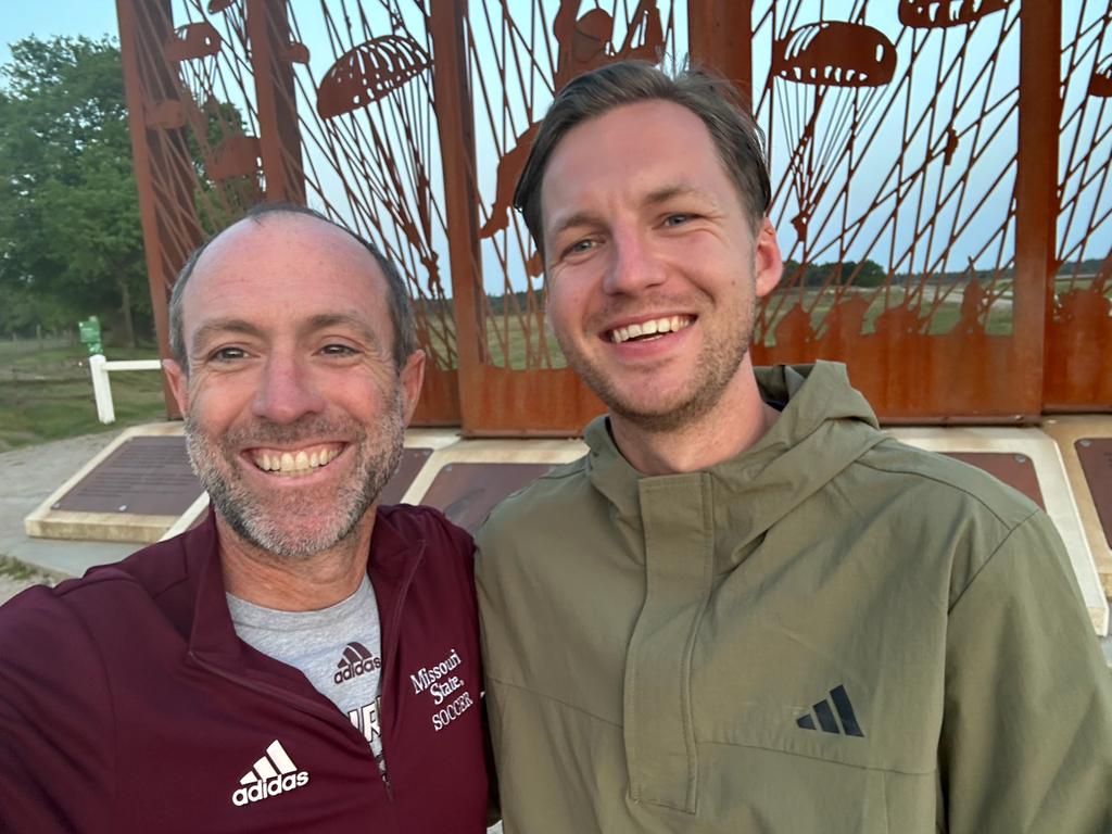 Two smiling men taking a selfie outdoors with a rust-colored decorative sculpture behind them.