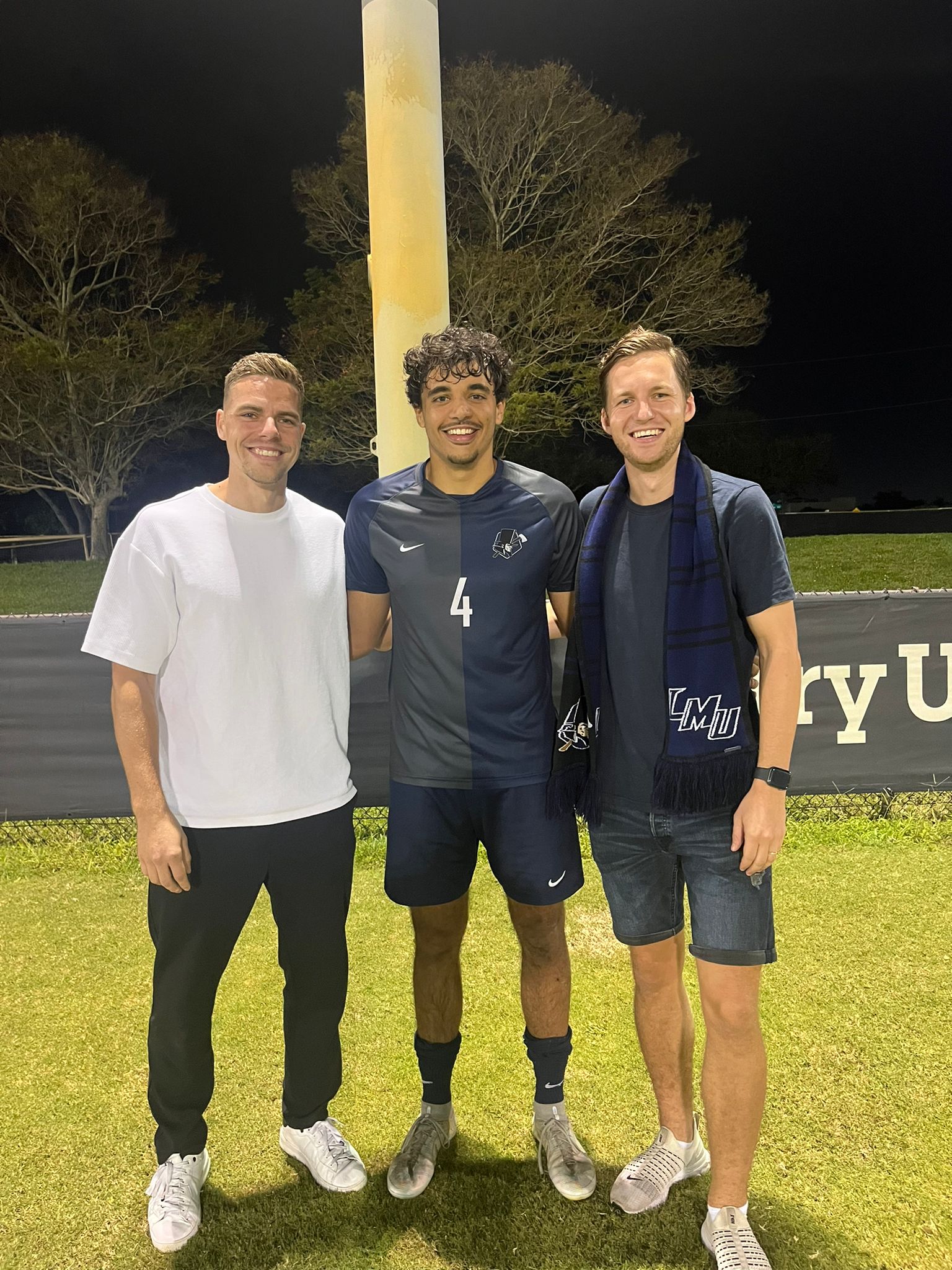 Three men smiling on a soccer field at night; the center one wears a number 4 soccer uniform.