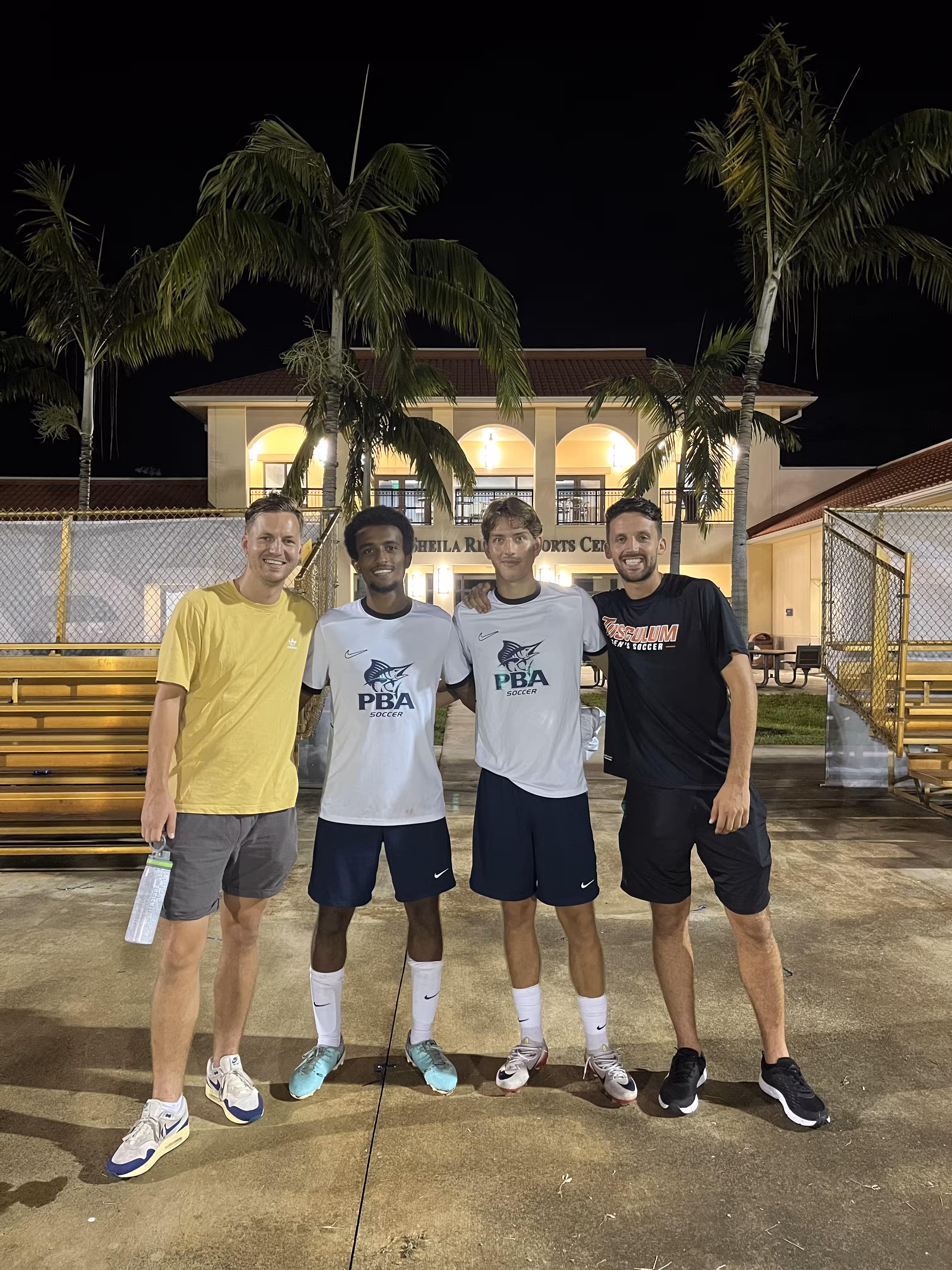 Four men posing at night in front of a sports center with palm trees and yellow benches.