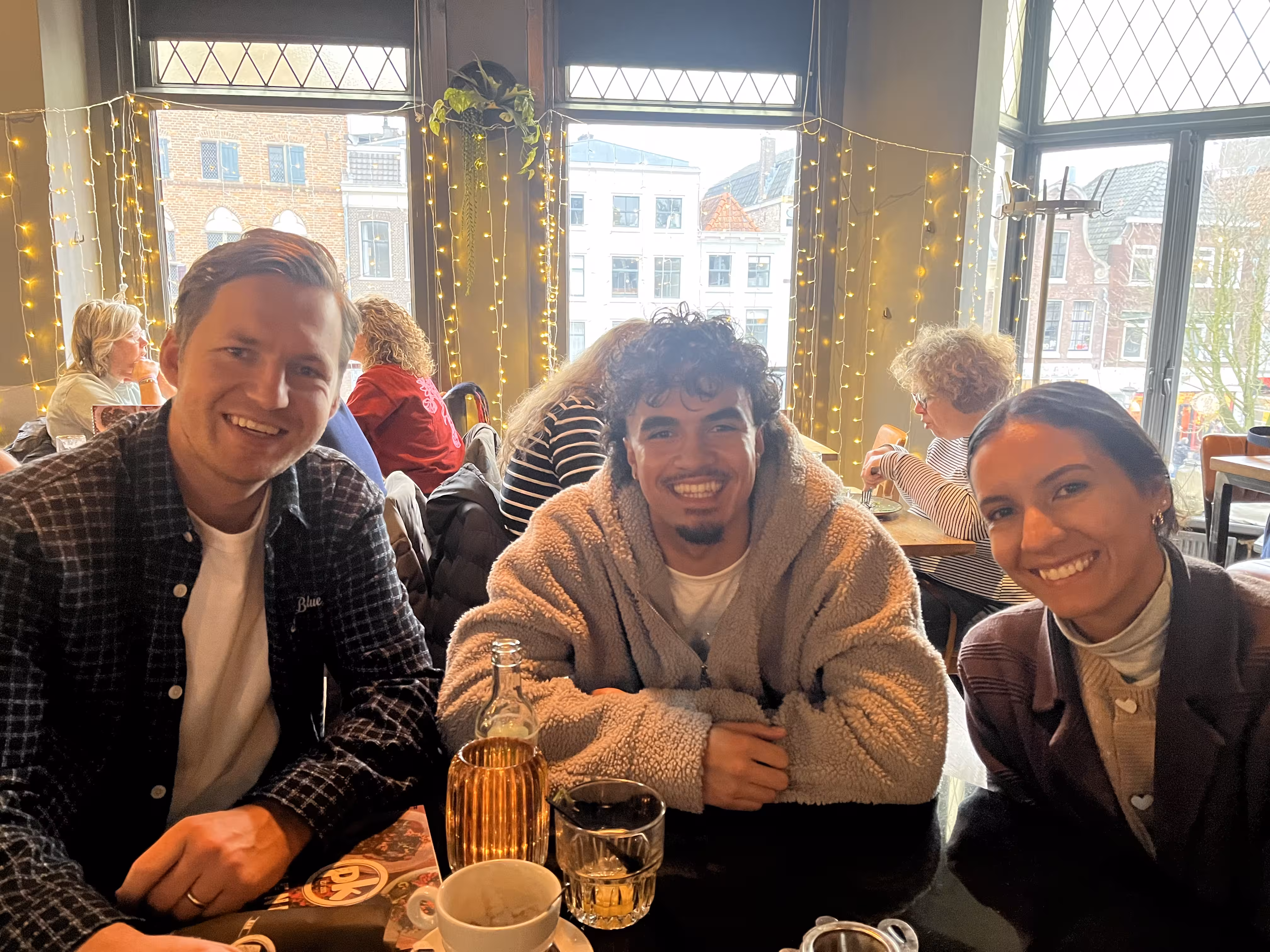 Three friends smiling at a cafe table with drinks, with string lights and large windows behind them.