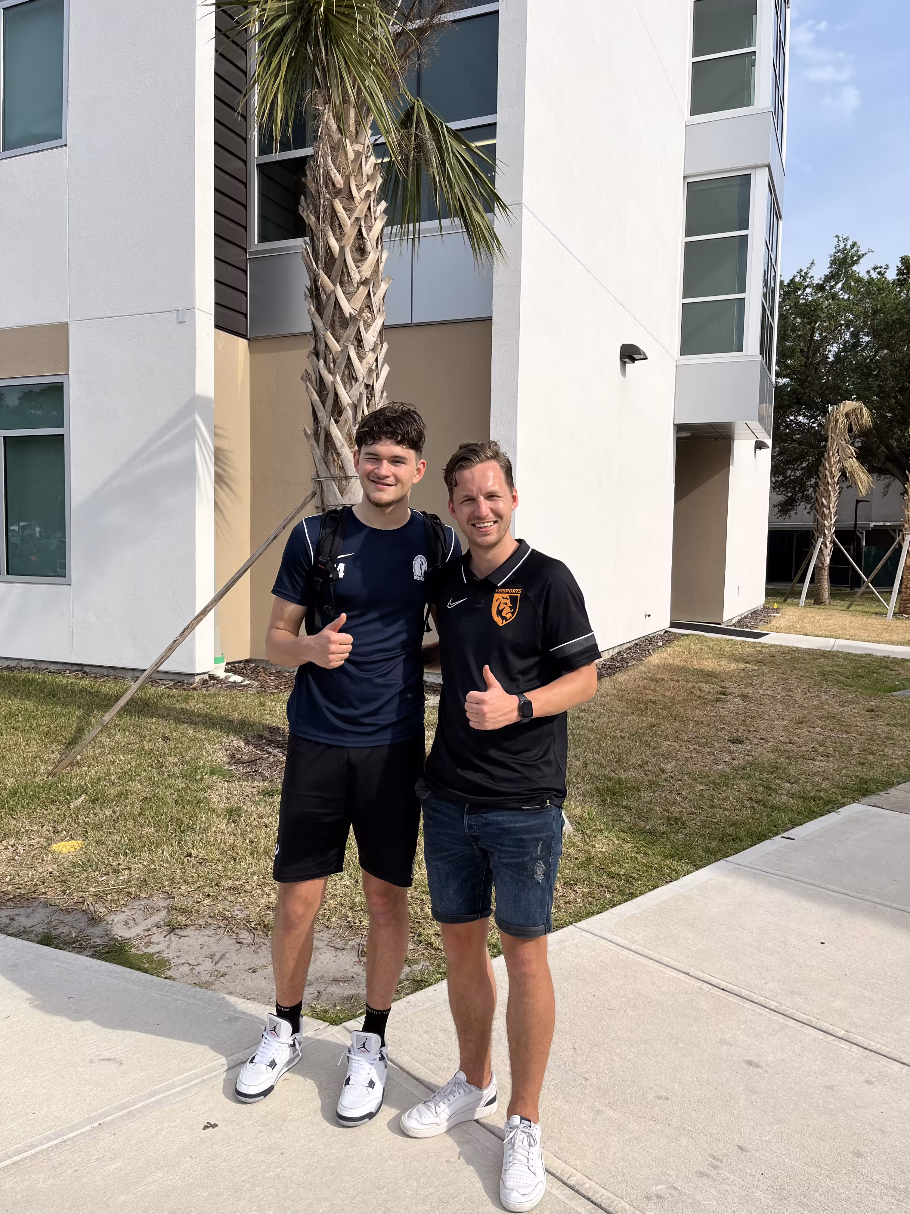 Two young men outdoors near a building and palm trees giving thumbs up and smiling at the camera.