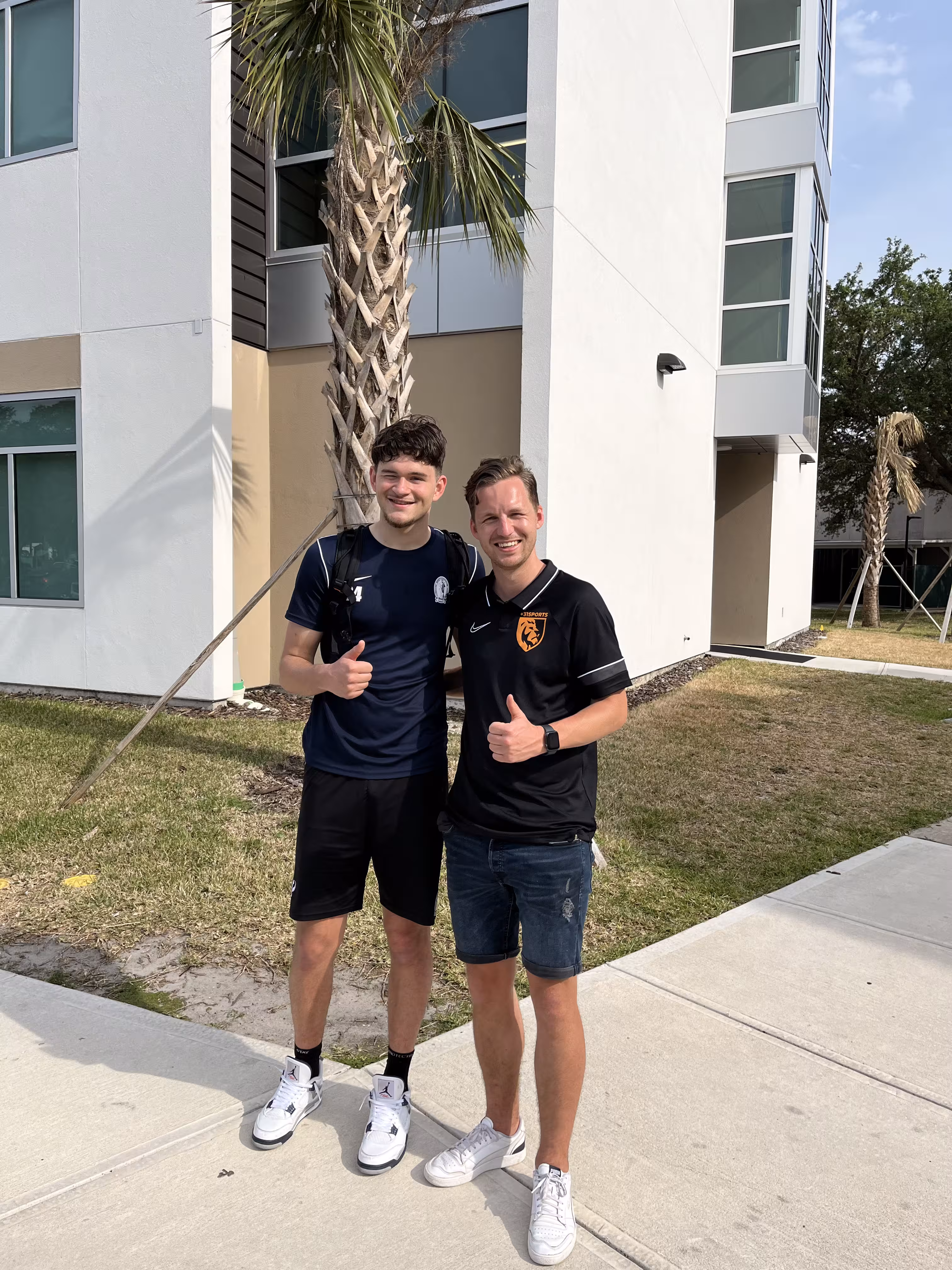 Two young men standing outside by a palm tree and building, smiling and giving thumbs up.