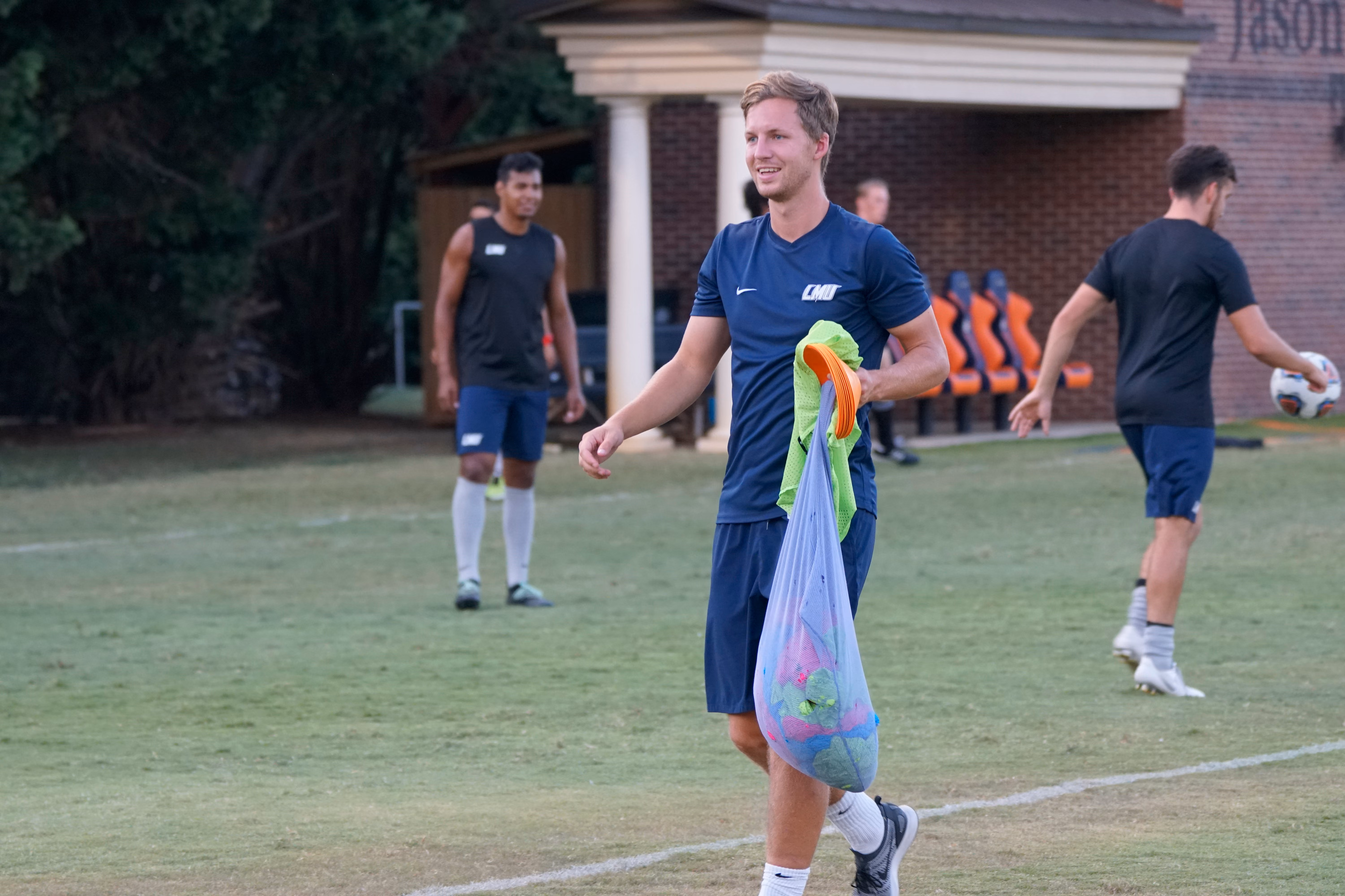 Man in blue sportswear carrying a bag of training cones walking on a soccer field.