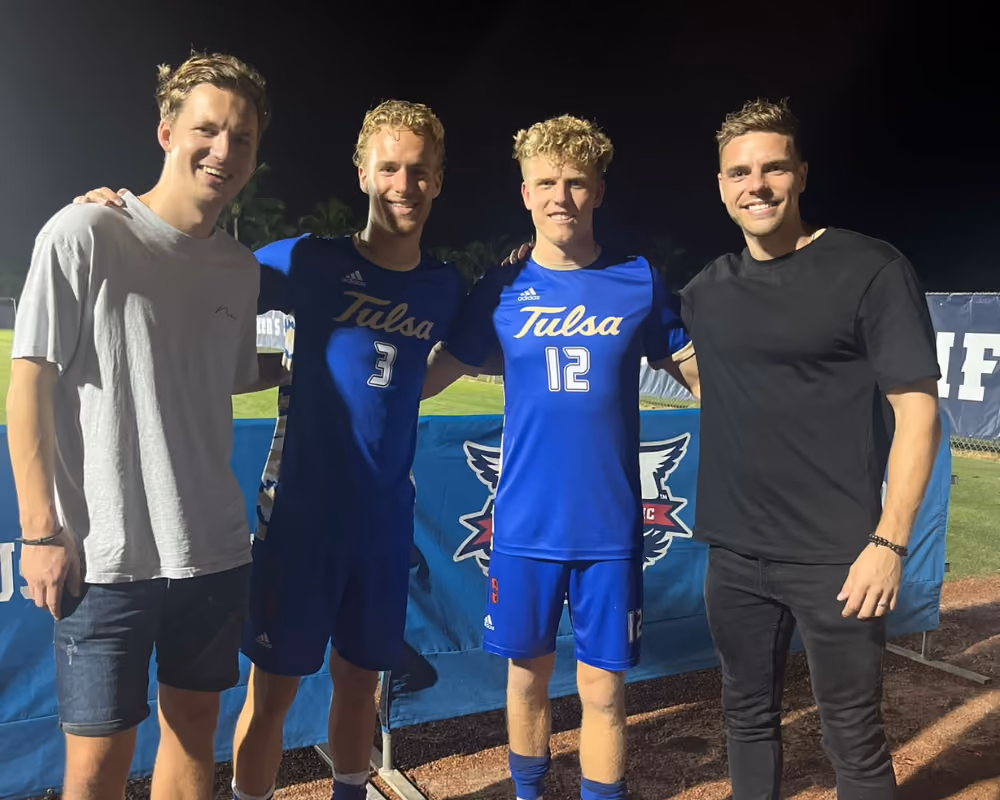 Four smiling men standing together at night, two wear blue Tulsa soccer uniforms numbered 3 and 12.