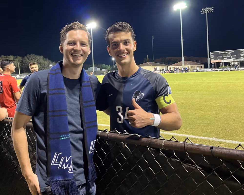 Two men smiling by a sports field at night, one wearing a soccer jersey with captain armband.