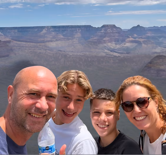Smiling family of four taking a selfie with canyon cliffs and sky in the background.