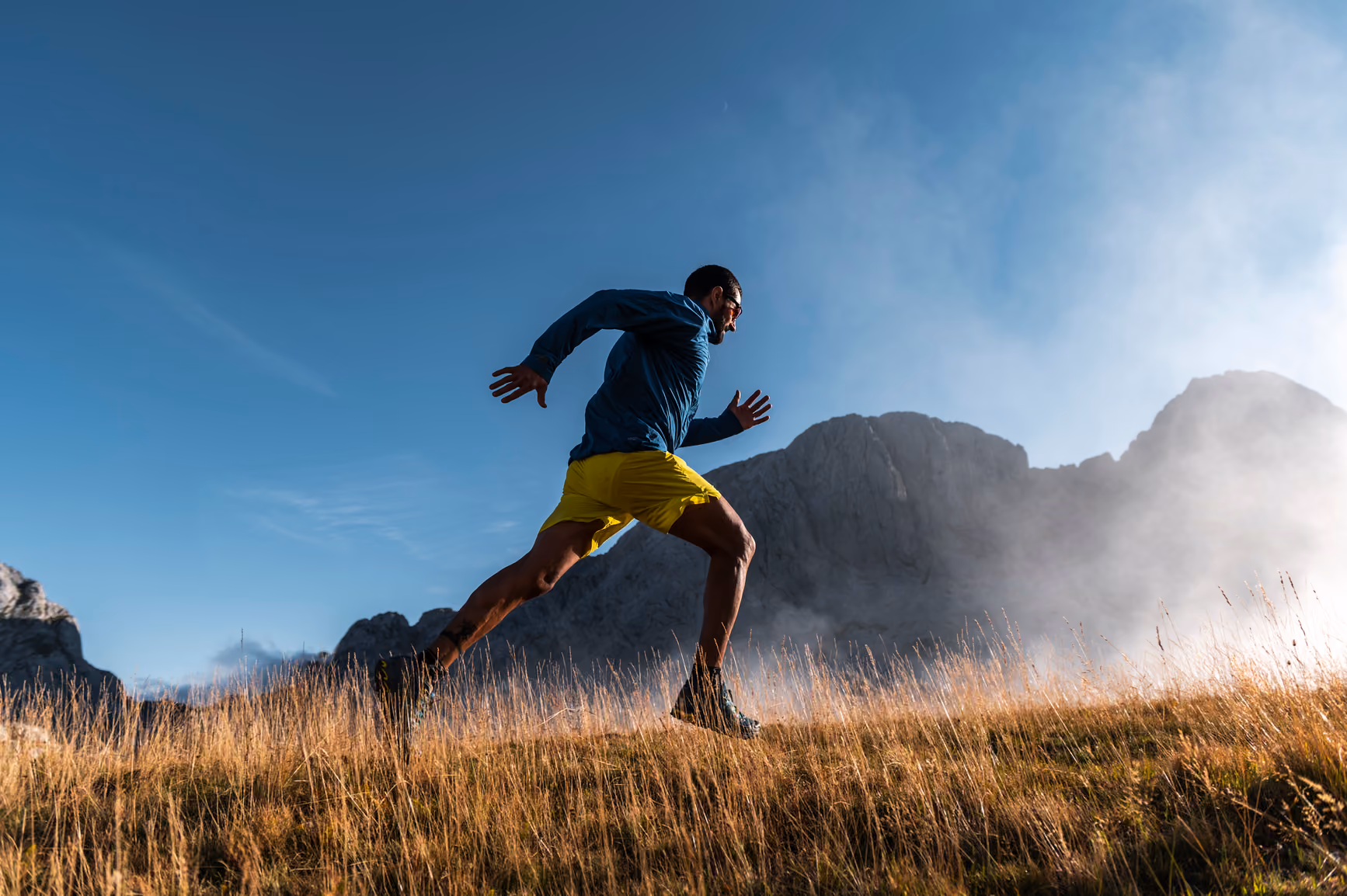 Man in yellow shorts running uphill on grassy terrain with mountains and blue sky in background.