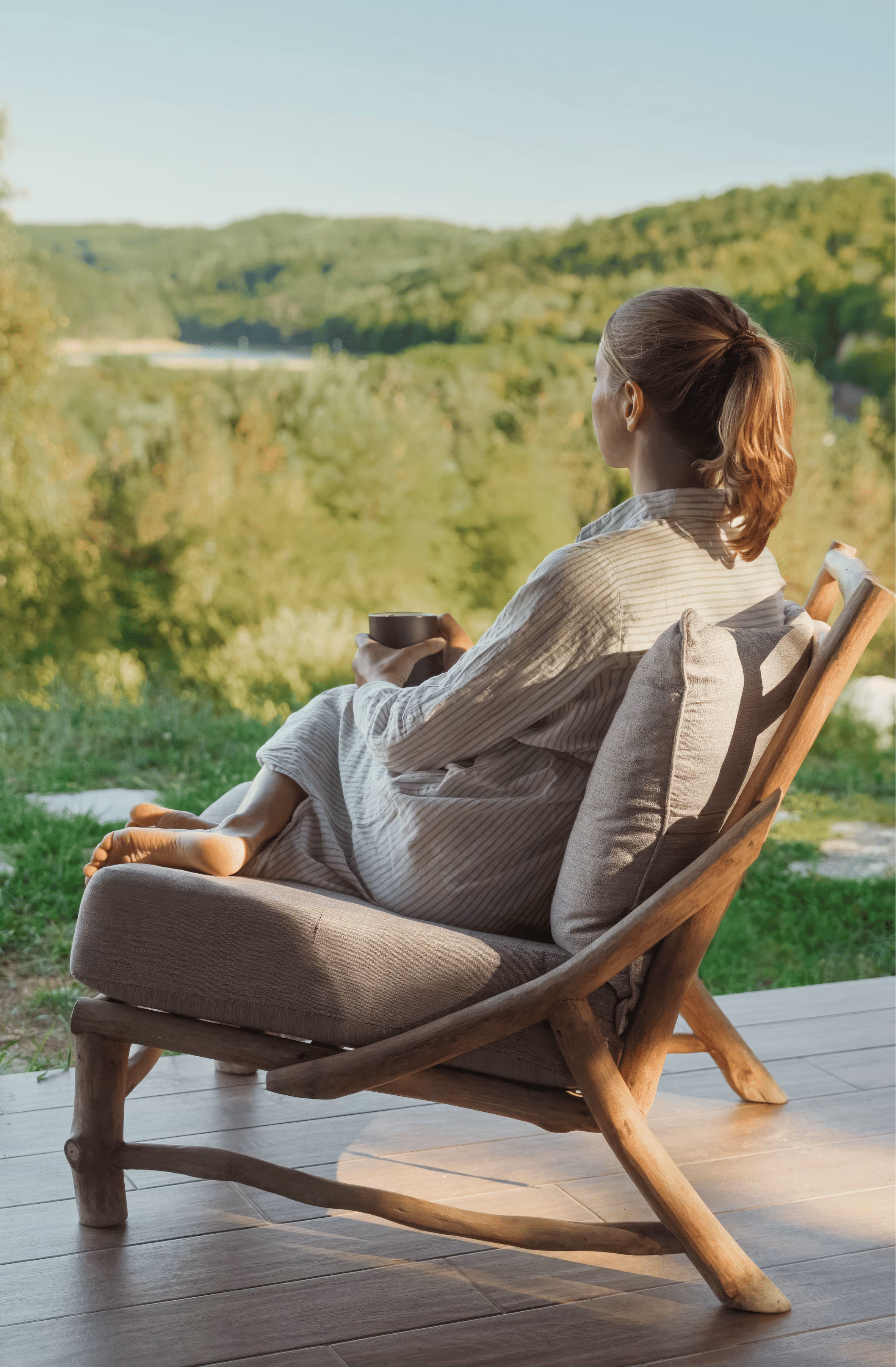Woman in a light gray robe sitting barefoot on a wooden lounge chair with cushions, holding a cup, facing a lush green landscape with trees and hills in soft sunlight.