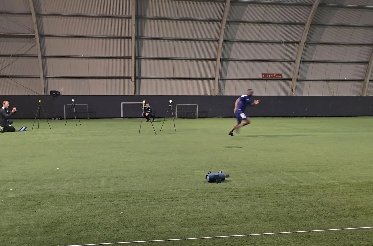 Maba Jarbou sprint over een indoor kunstgrasveld tijdens een snelheidstest, begeleiders volgen met meetapparatuur op statieven.