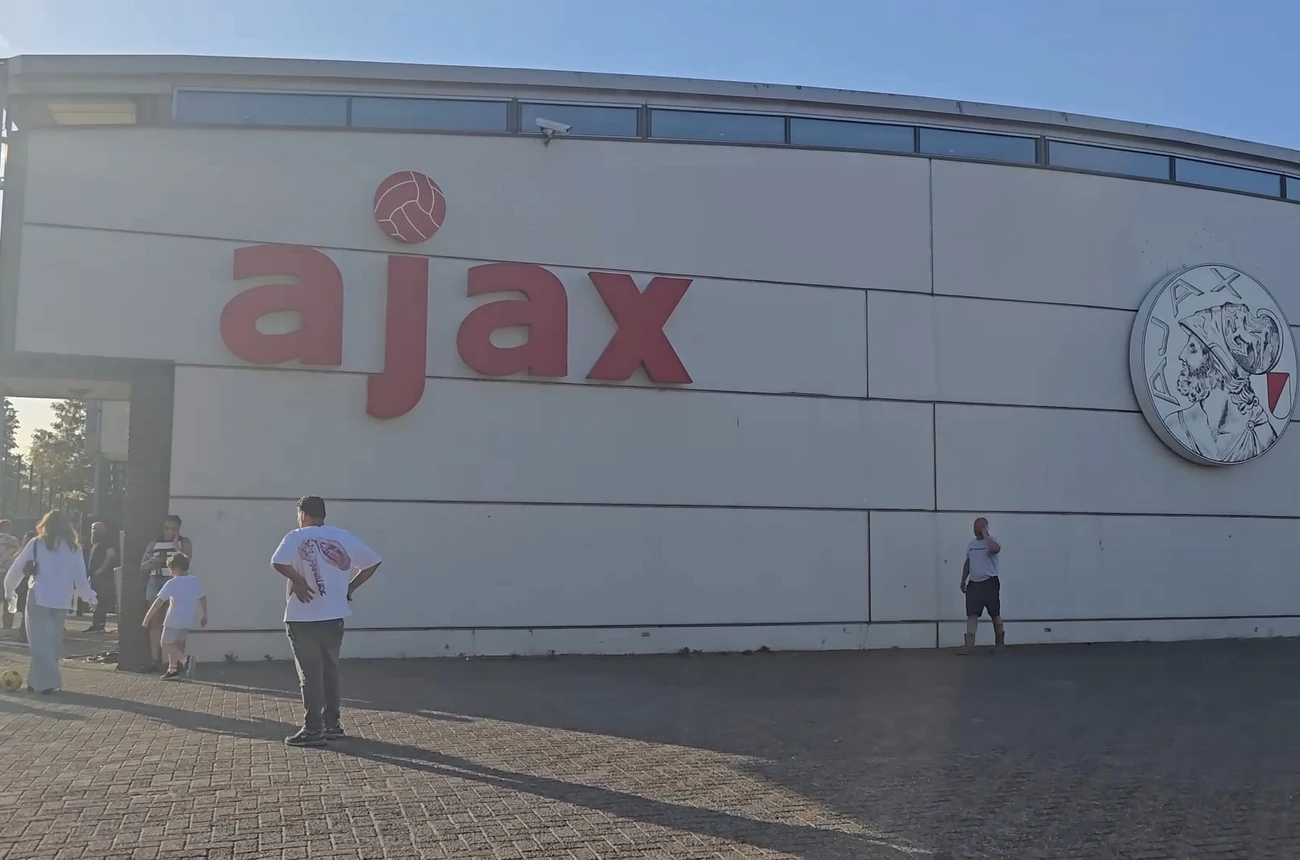 Buitengevel van het Ajax-complex de Toekomst met het Ajax-logo, mensen lopen voorbij in de zon.