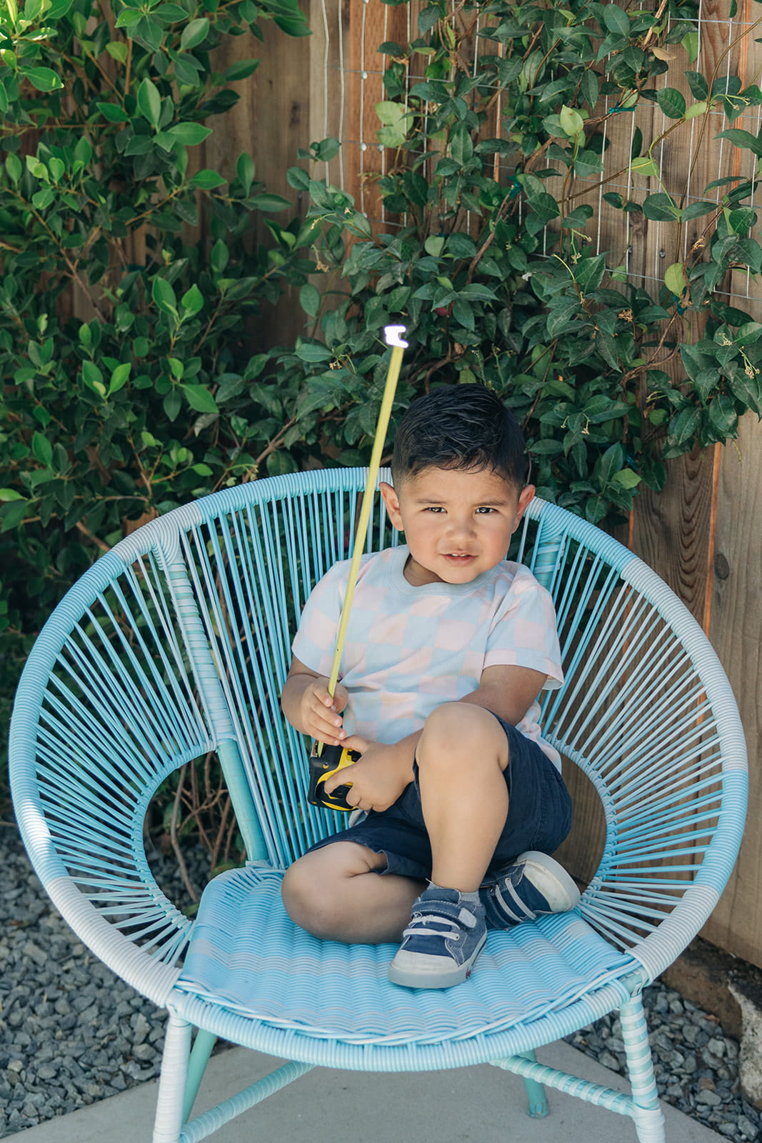 Young boy sitting on a blue woven chair outdoors holding a yellow measuring tape.