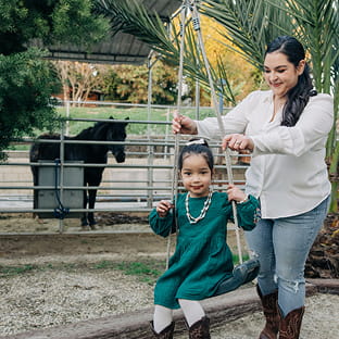 Alejandra (Ale) Aviña pushes a smiling girl on a swing near a palm tree with a horse in a fenced area behind them.