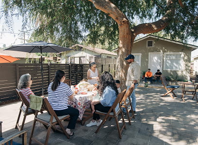 Five people gathered outdoors around a table with food under a tree, two more seated in the background.