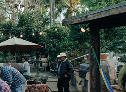 People working outdoors in a garden area with tools, umbrellas, and greenery in the background.