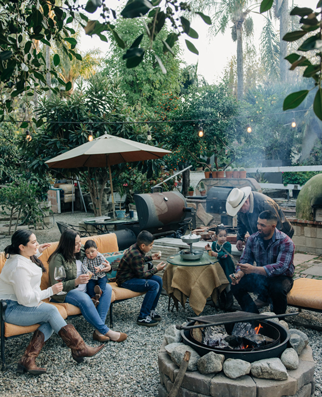 Family gathered around a fire pit in a garden, roasting food and sitting on benches with drinks.