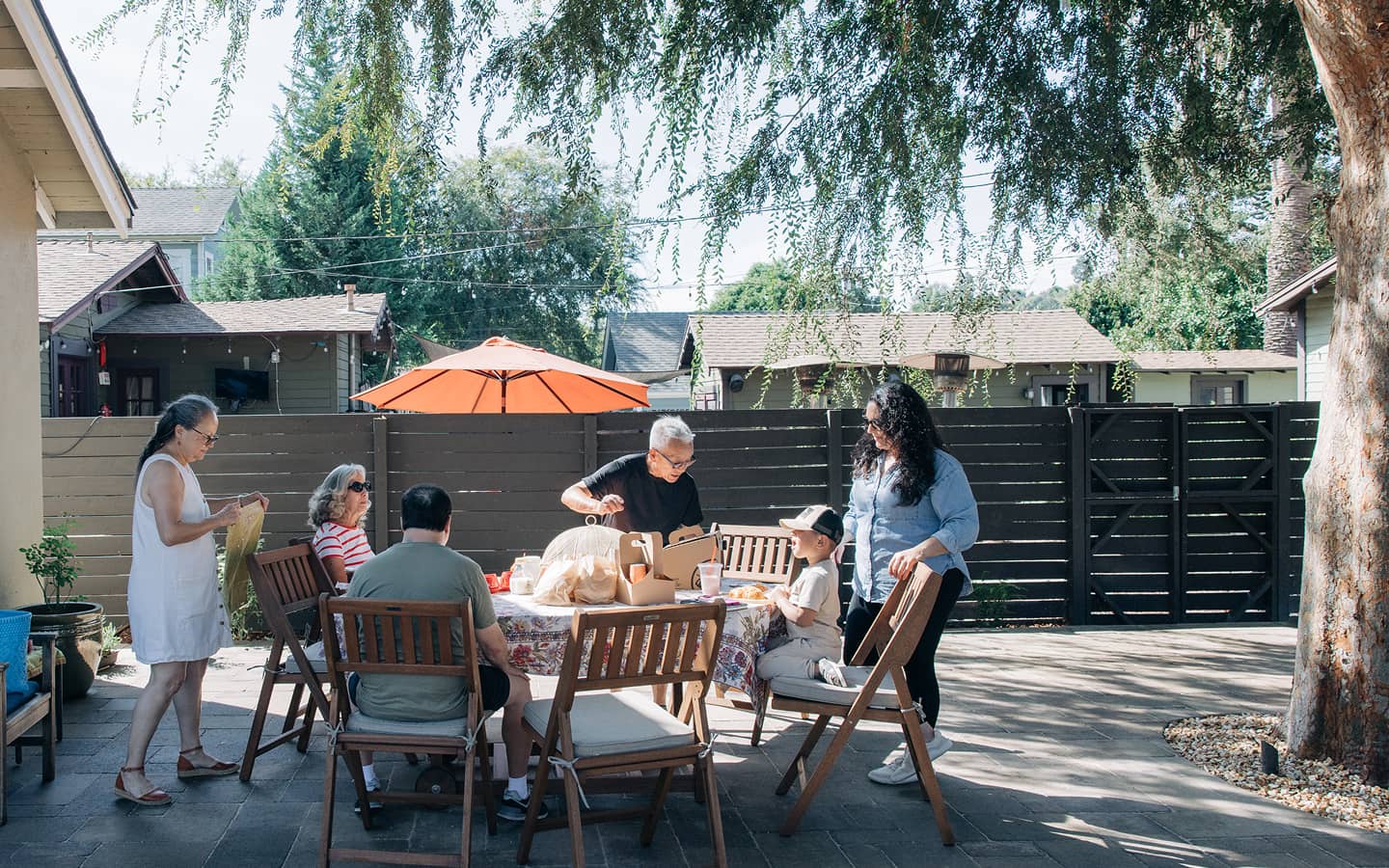 Family gathers around a table with food under a tree in a backyard on a sunny day.