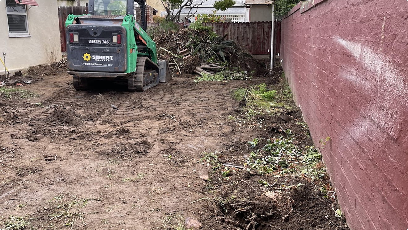 Green skid steer loader in a dirt backyard clearing debris near a wooden fence and red brick wall.