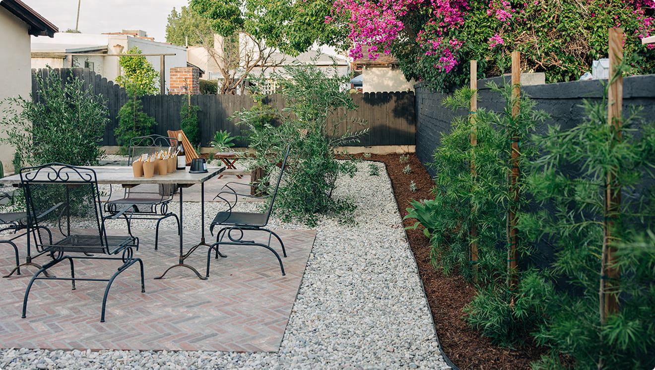 Outdoor patio with metal chairs and table on brick and gravel, surrounded by greenery and a flowering tree.