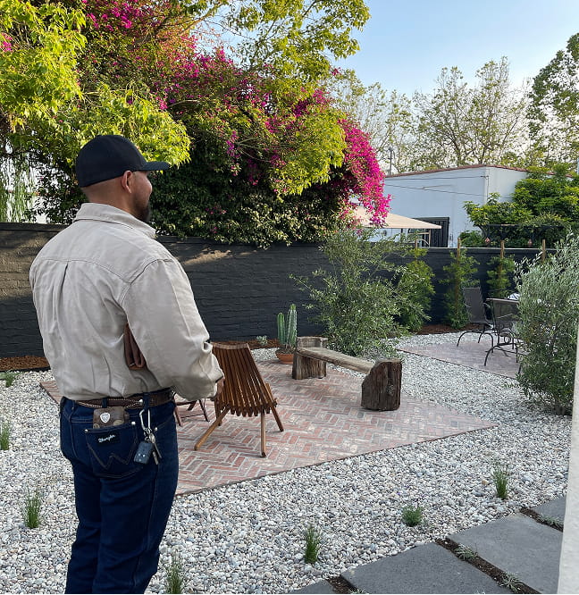 Jonathan Aviña, in a cap and jacket, stands in a landscaped backyard with gravel, chairs, and flowering shrubs.