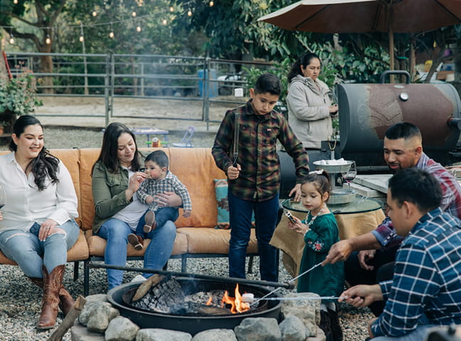 Family gathered outdoors around fire pit roasting marshmallows, with a barbecue grill nearby.