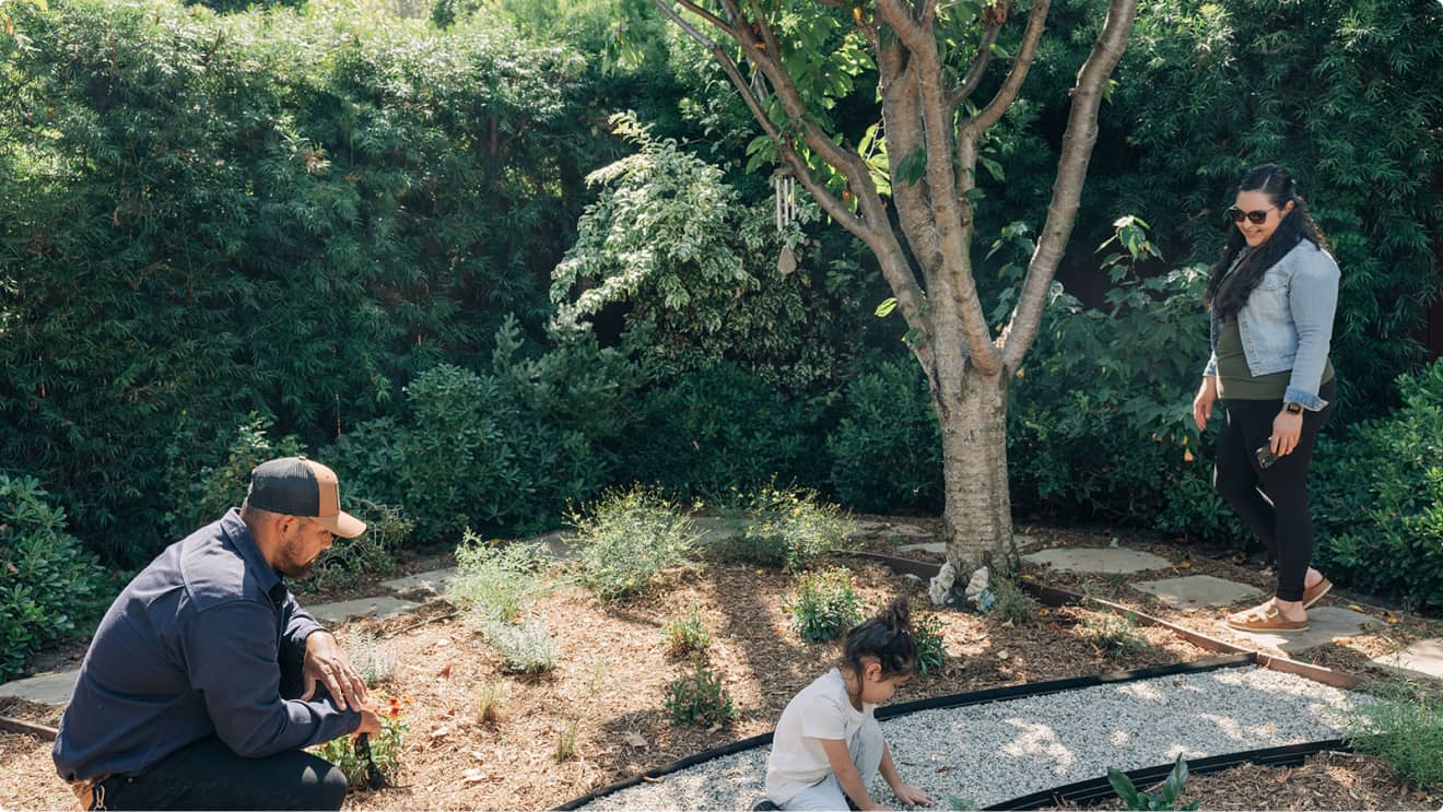 A man crouches by plants, a child plays on gravel, and a woman watches near a tree in a garden.