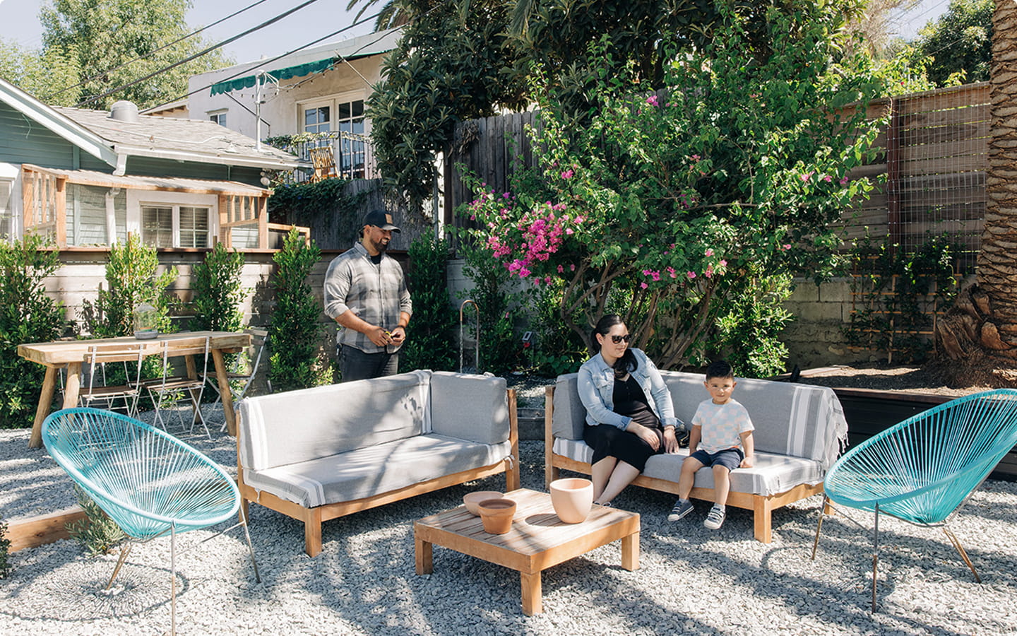 Man standing and woman with child sitting on outdoor sofas in a backyard with greenery and blue chairs.