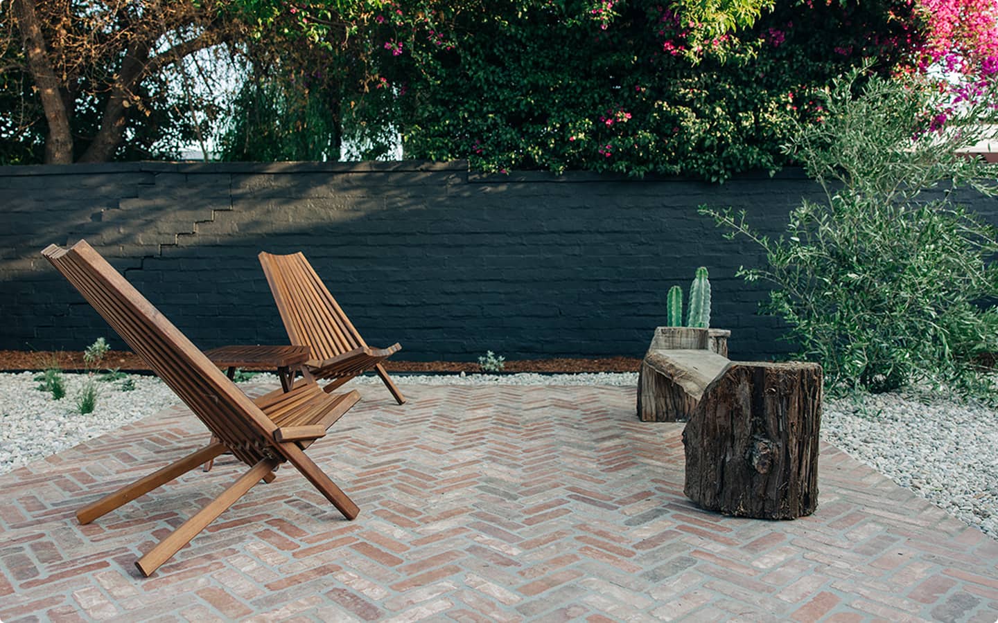 Wooden chairs and a log bench on a herringbone brick patio surrounded by gravel and plants.