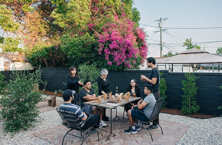 Group of seven people gathered outdoors around a table with drinks, chess set, and snacks.