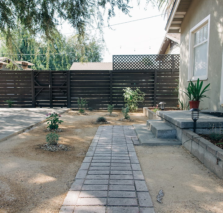 Yard with a paved walkway, small plants, stairs leading to a house with a large window and fence.