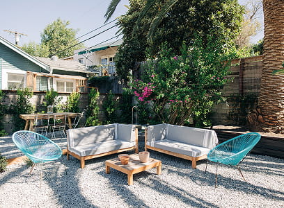 Outdoor patio with two gray couches, two blue chairs, and a wooden coffee table on gravel.