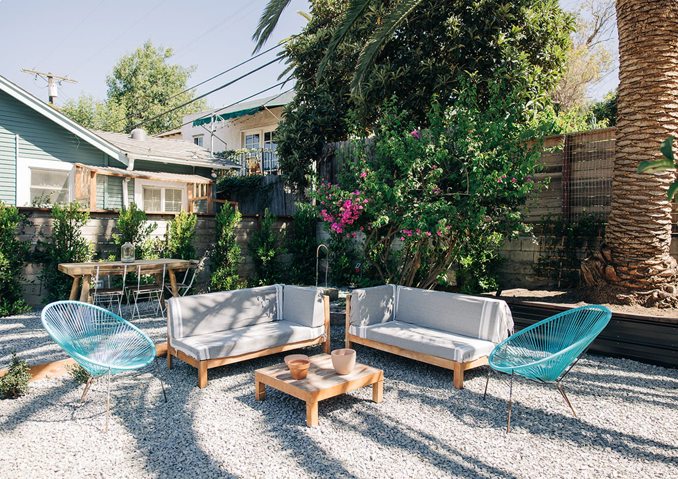 Outdoor seating area with two gray sofas, two blue chairs, a wooden table, and greenery in background.