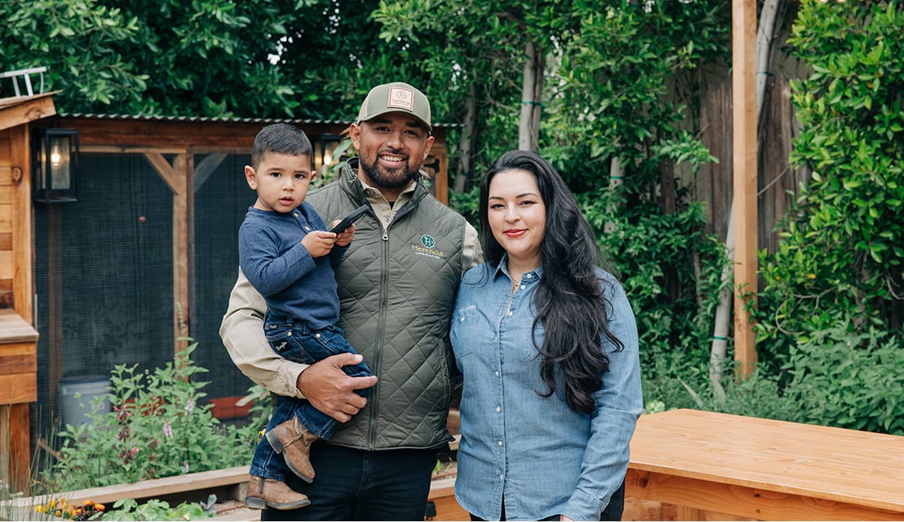 Man holding a child and woman standing together outdoors with garden and wooden structure in background.