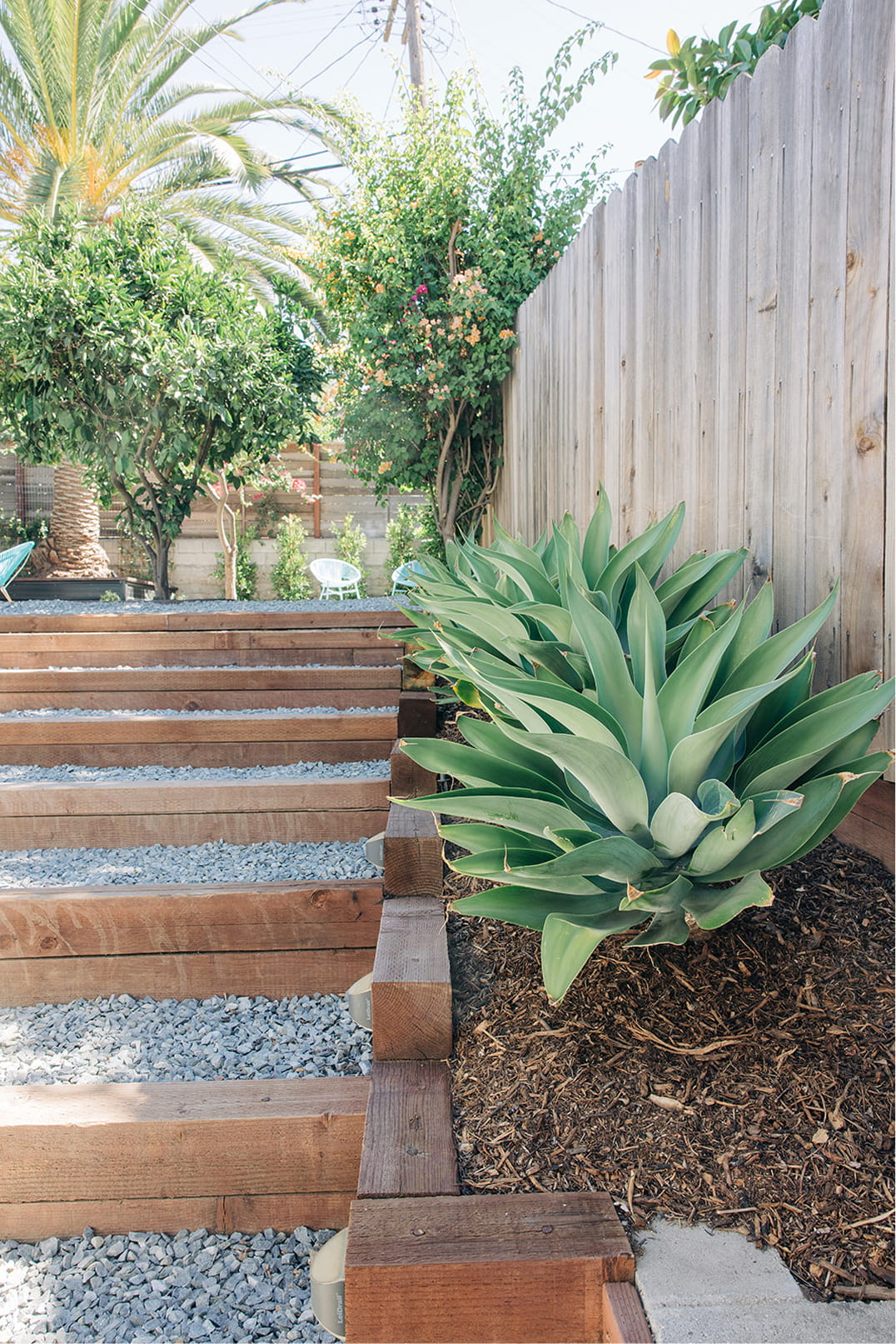 Wooden steps with gravel next to a large succulent and a wooden fence in a landscaped backyard.