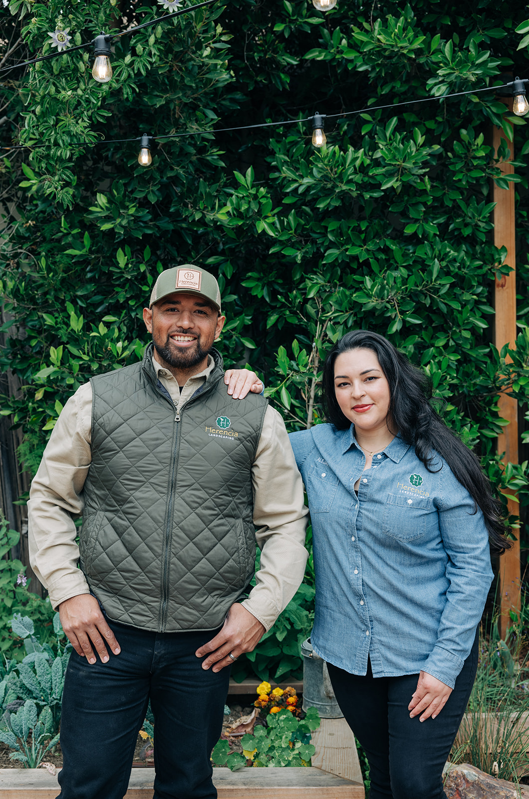 Man and woman standing outdoors with greenery and string lights, wearing Herencia Landscaping shirts.