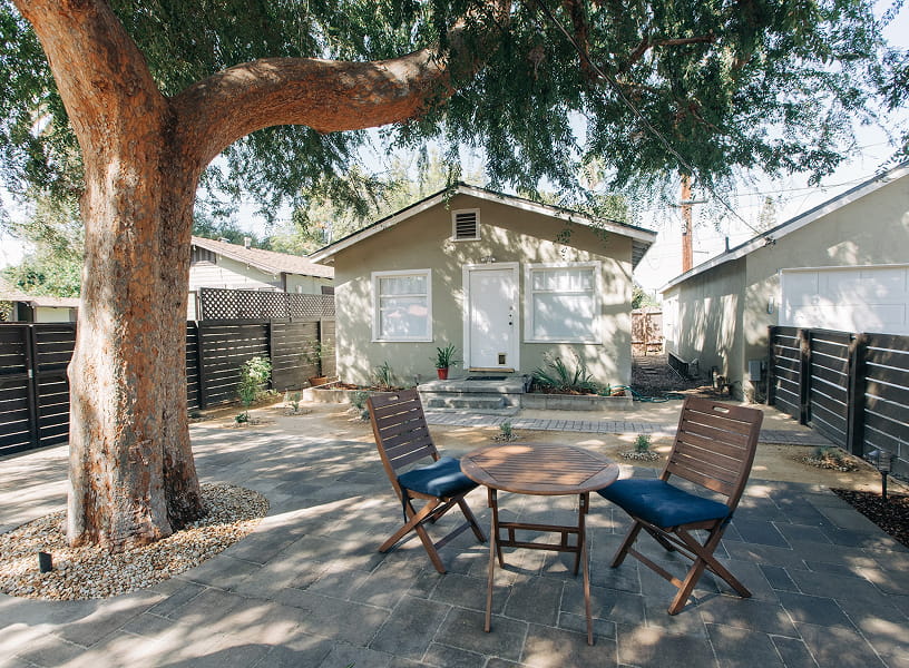 Backyard patio with a large tree, two wooden chairs with blue cushions, and a round table in front of a small house.