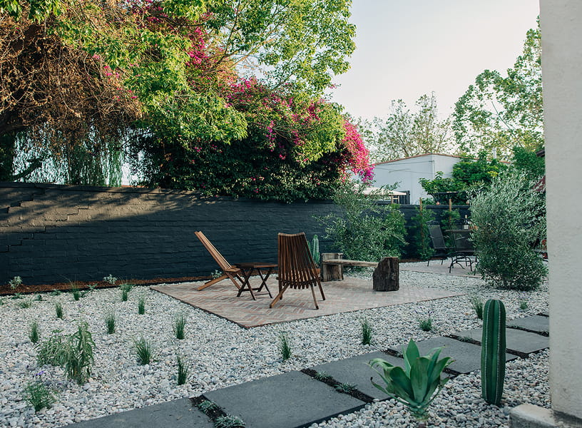 Minimalist backyard with stone patio, wooden chairs, small plants, and black brick wall.