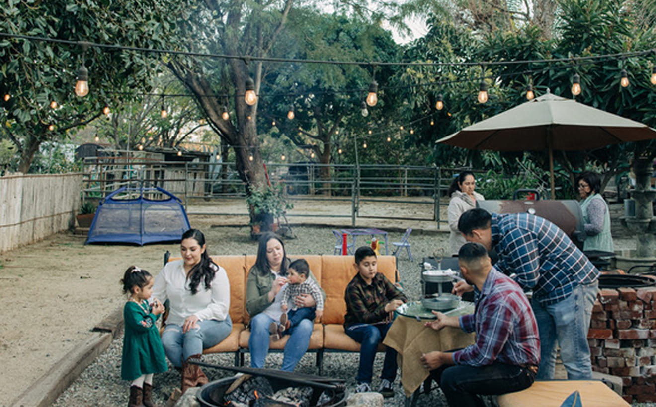 Family gathered outdoors around a fire pit with string lights and a grill in the background.