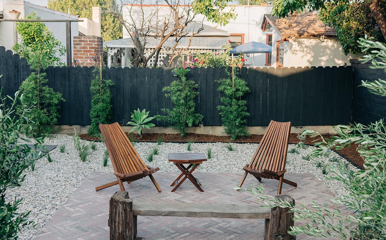 Backyard patio with two wooden chairs and small table on brick patio surrounded by plants and black fence.