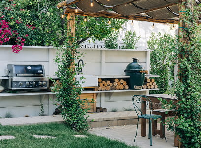 Outdoor kitchen with grill, green ceramic smoker, wood storage, and a metal chair under a pergola.