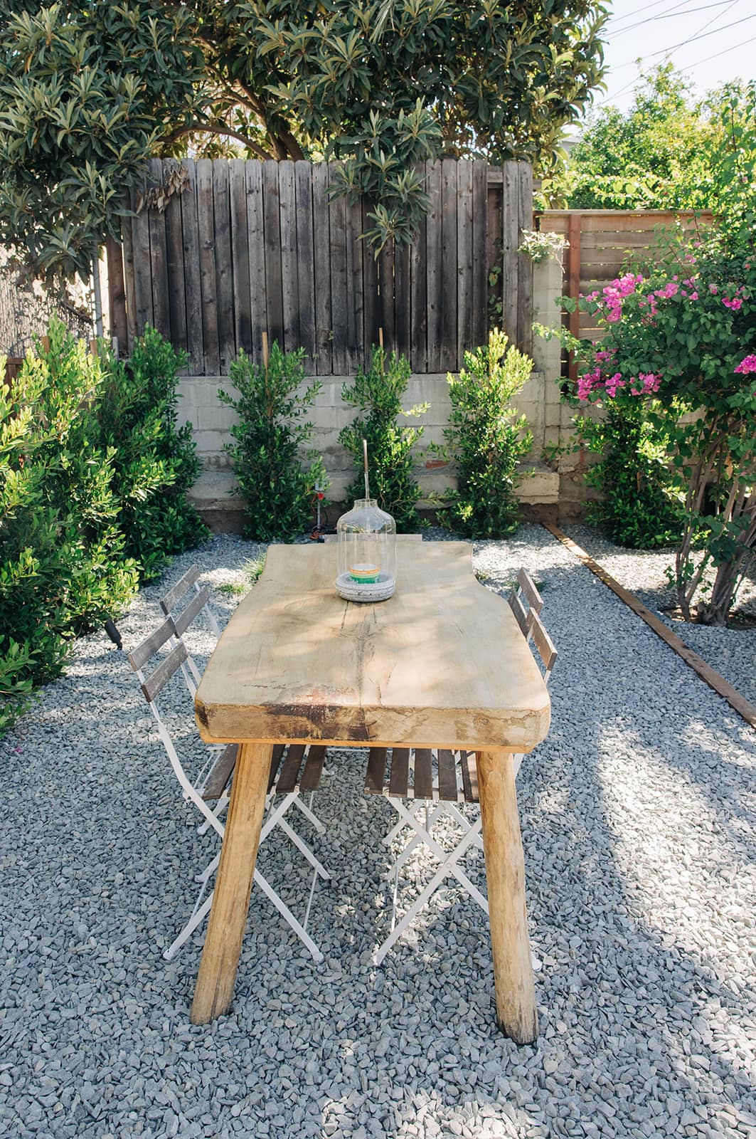 Outdoor wooden table with four metal chairs on gravel beside green shrubs and pink flowers.