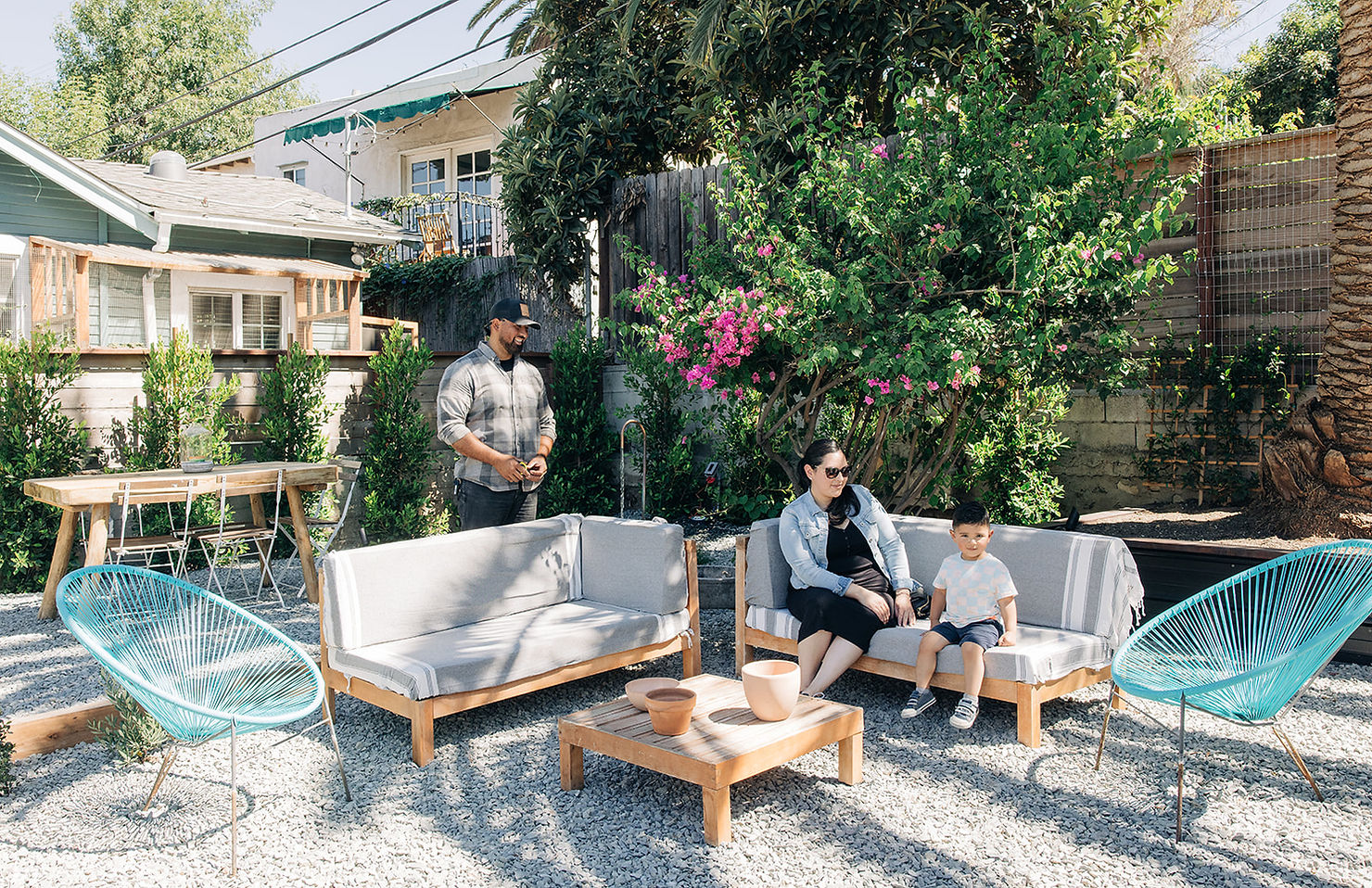 Man standing, woman and child sitting on outdoor sofa, sunny garden with green plants and pink flowers, teal chairs, relaxed mood.