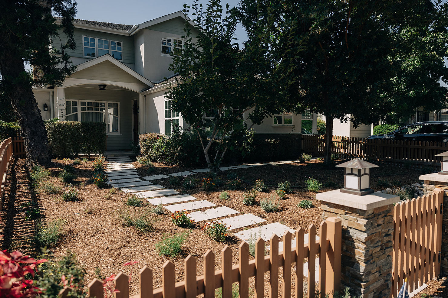 Two-story house with green and beige exterior, surrounded by a garden with stepping stones. Brown picket fence, trees, and a black car.