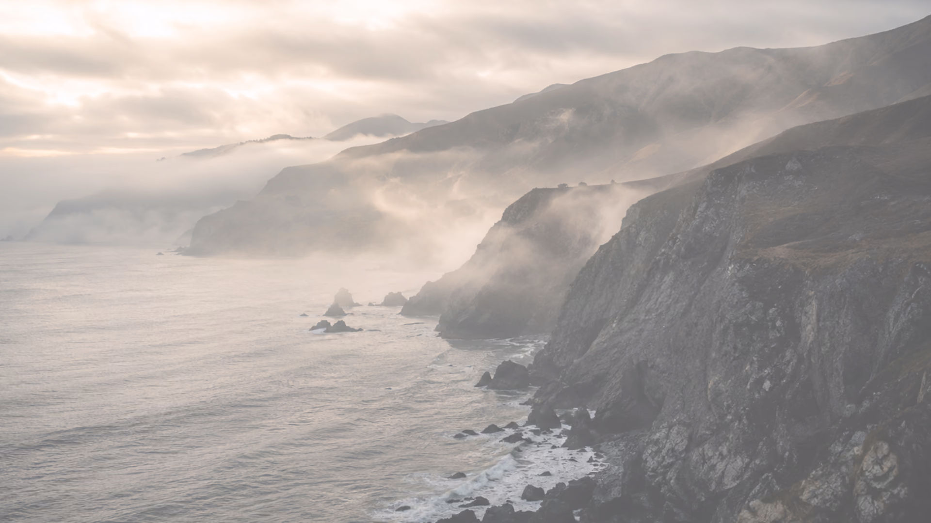 Foggy coastal cliffs with waves breaking on rocks under a cloudy sky at sunrise or sunset.