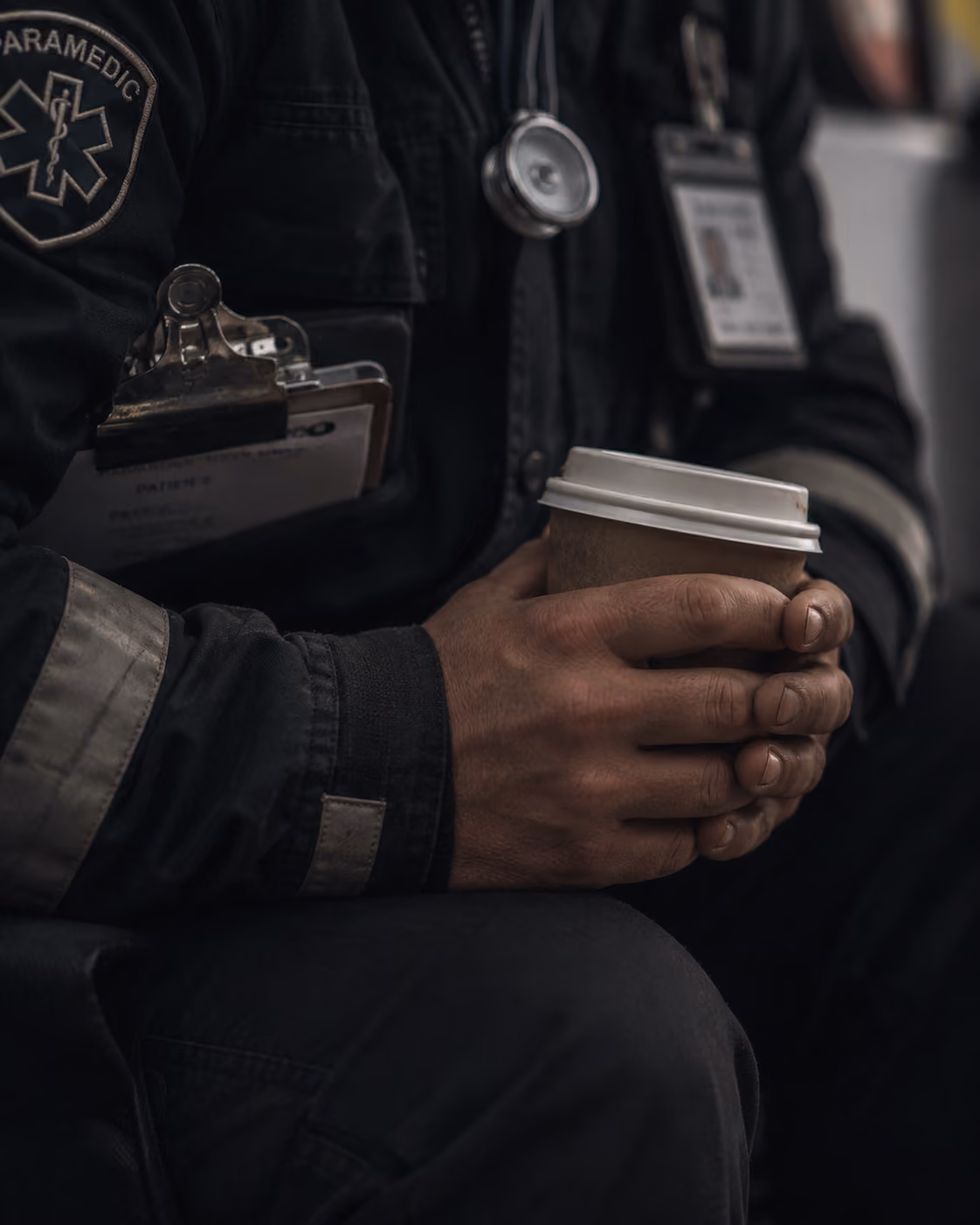 Paramedic in uniform holding a paper coffee cup with both hands, showing a badge and clipboard.