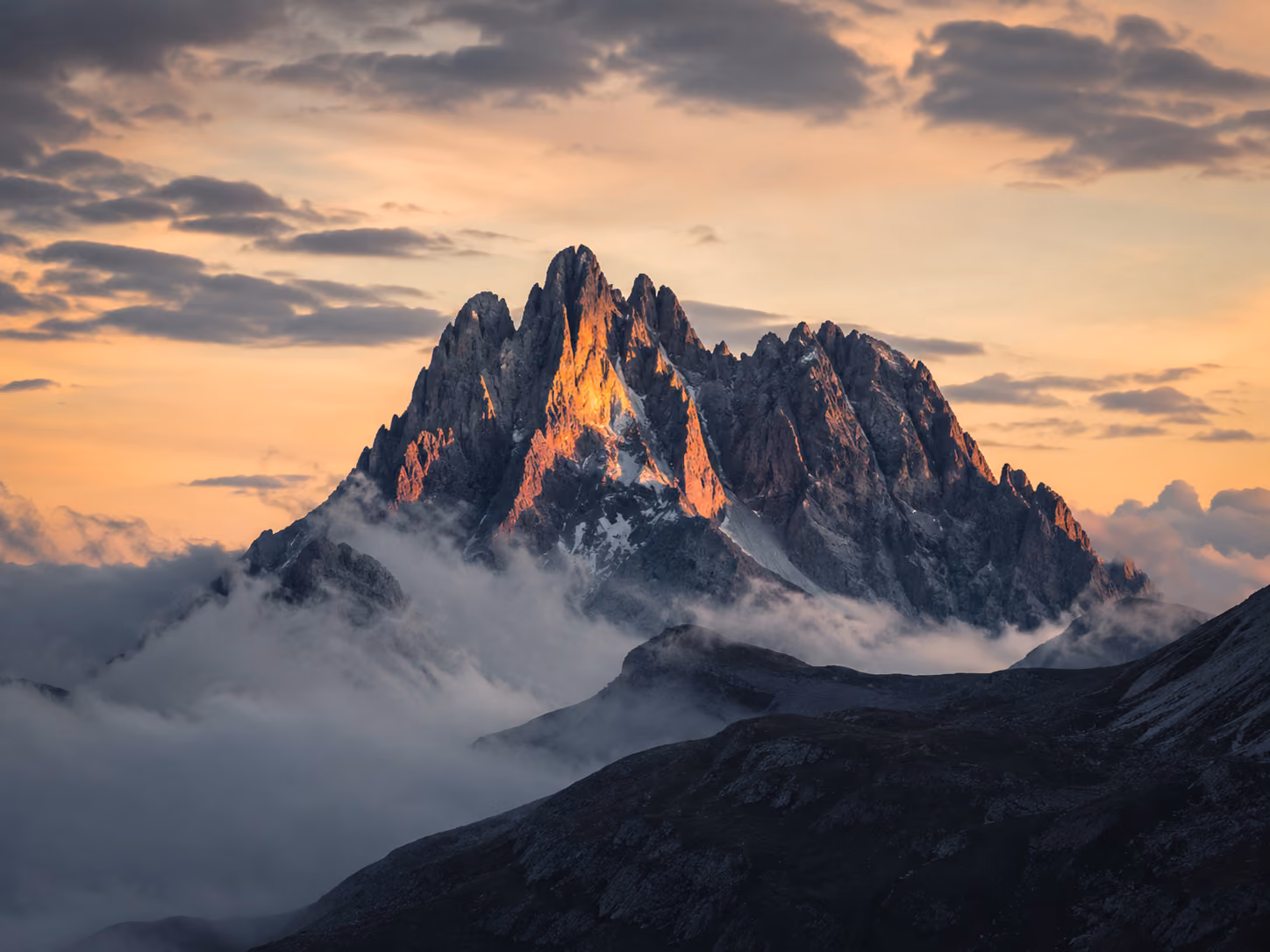 Jagged mountain peak lit by warm sunset light above low clouds under a partly cloudy sky.