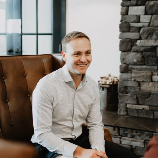 Smiling man in a white shirt sitting on a brown leather chair near a stone wall fireplace.