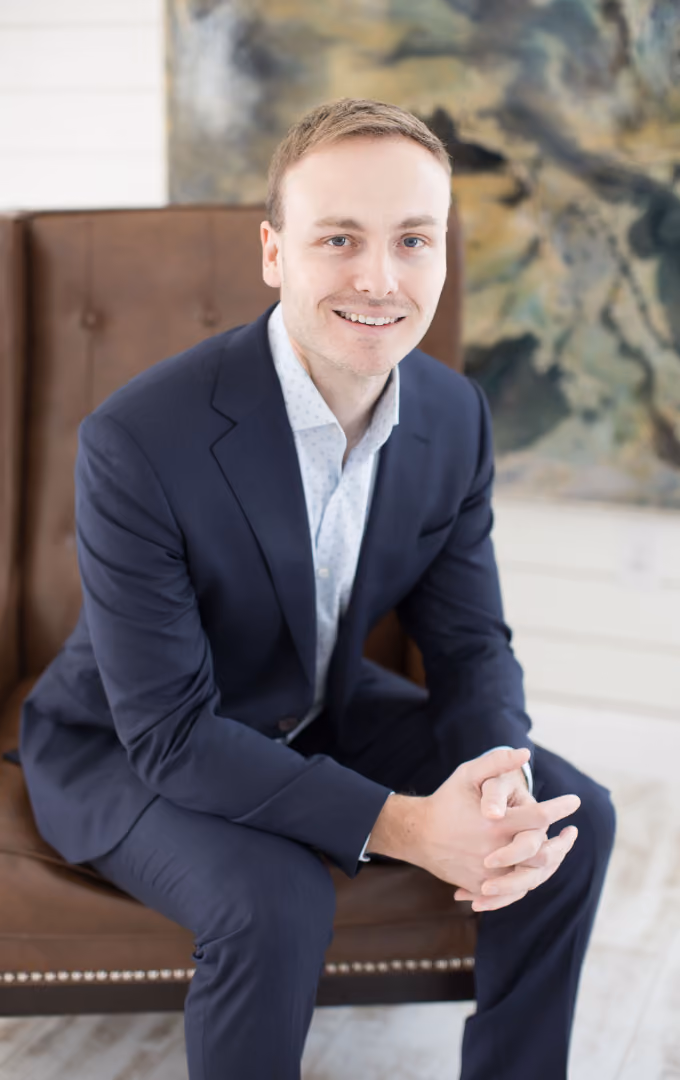 Young man in a navy suit sitting on a brown chair smiling with hands clasped.
